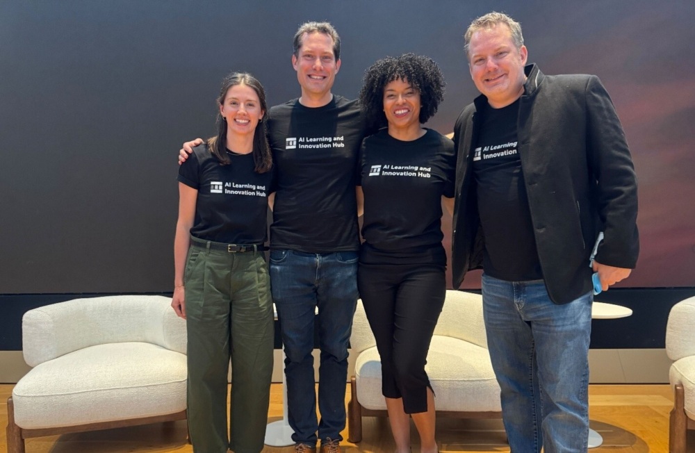 Four people stand side by side, smiling, all wearing matching black "AI Learning and Innovation Hub" t-shirts, in a modern indoor setting with armchairs and a wooden floor.